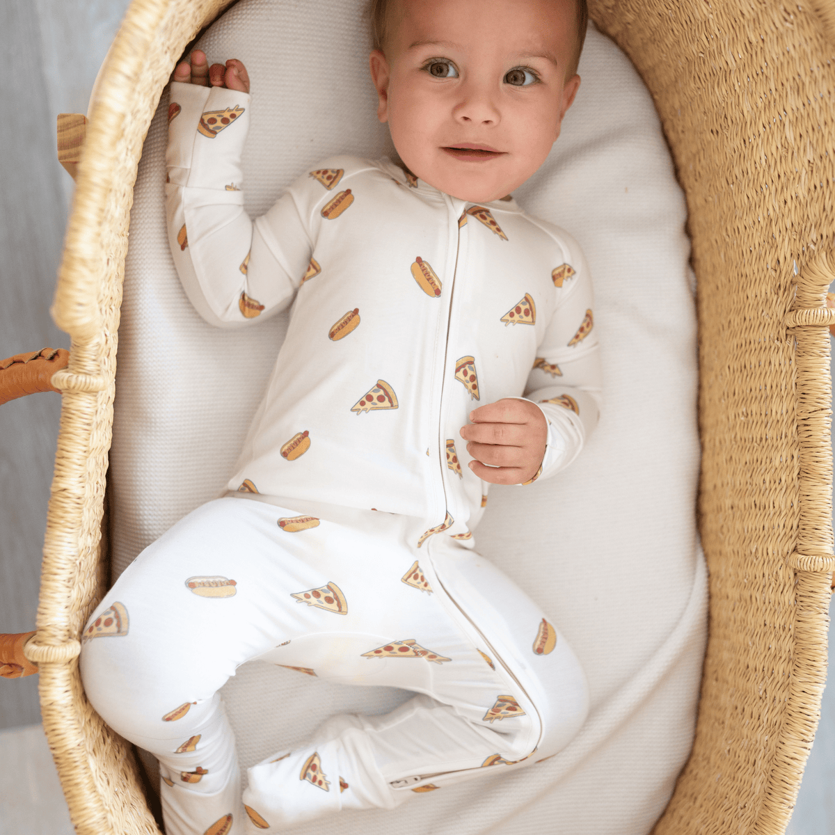 Baby in a white romper with pizzas and hot dogs  sitting in a wicker crib.