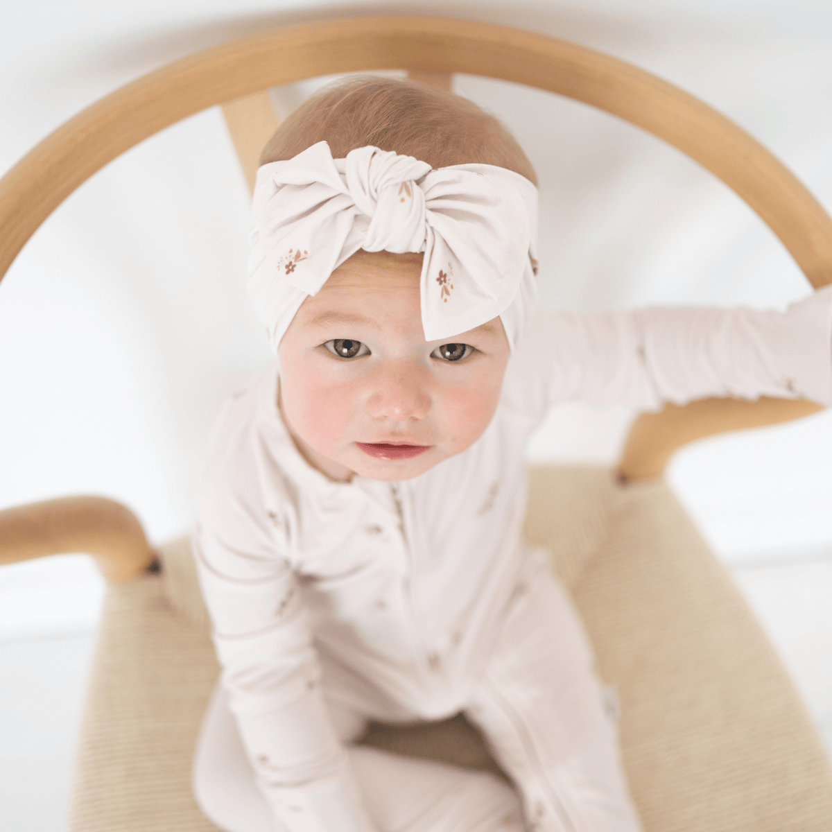Baby wearing a pink headband with a bow, sitting in a wooden chair.
