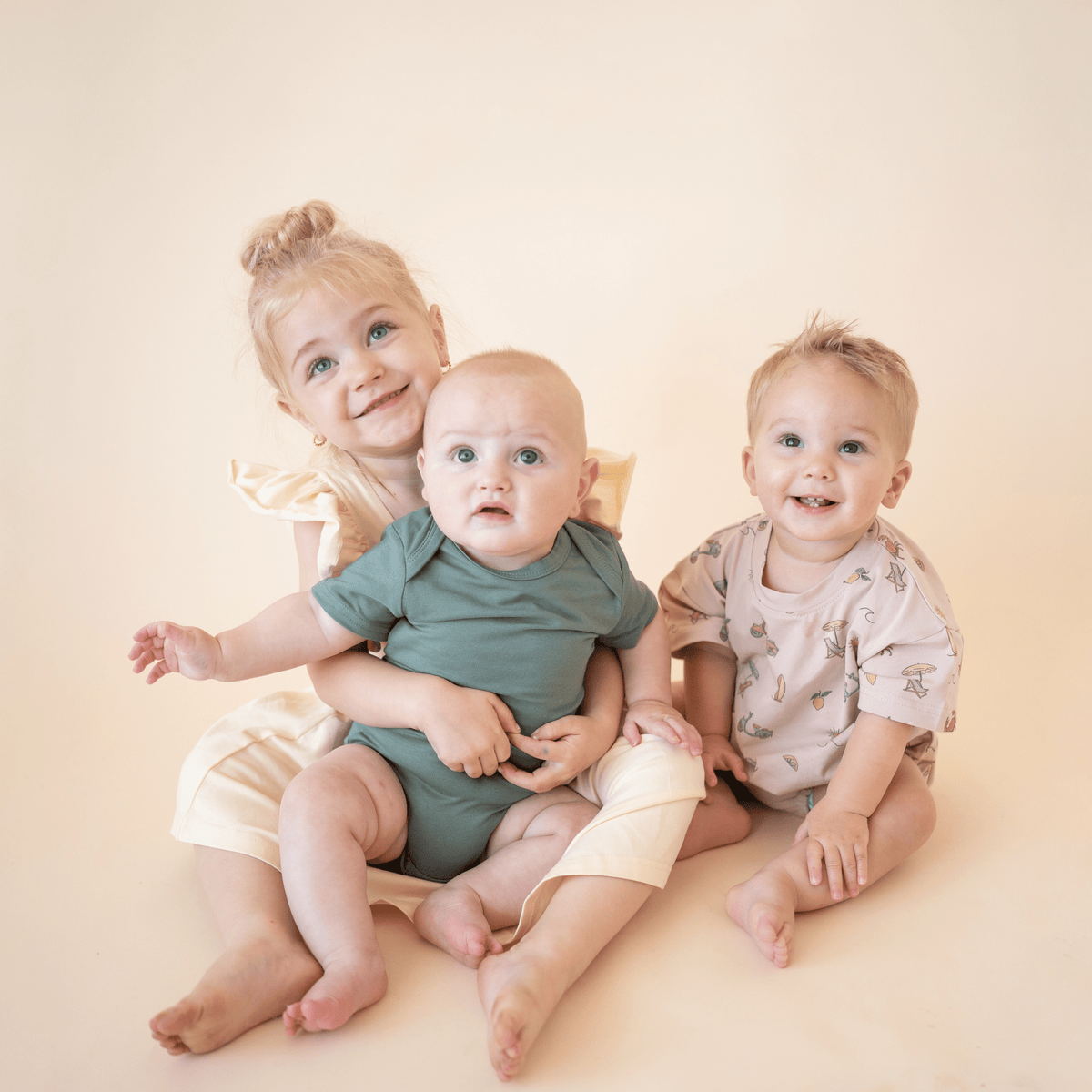 Three young children wearing bug + bean organic cotton outfits in coordinating summer colors, including a yellow dress, sage green bodysuit, and Summer Vibes printed romper