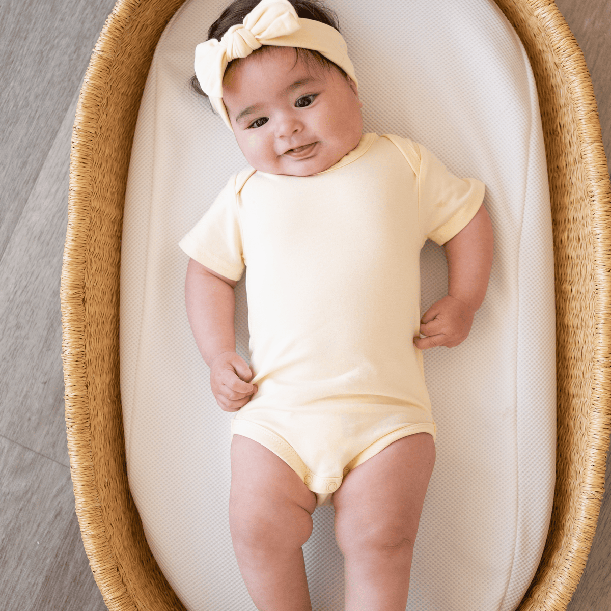Smiling baby lying in a Moses basket wearing the organic cotton Lemonade Yellow bodysuit and matching headband from bug + bean kids