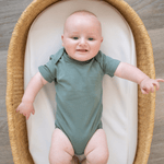 Baby wearing sage green organic cotton bodysuit, lying in a woven basket on a soft white mattress