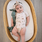 Baby girl lying in basket wearing Summer Vibes print short sleeve bodysuit with matching bow and holding a green bunny lovey