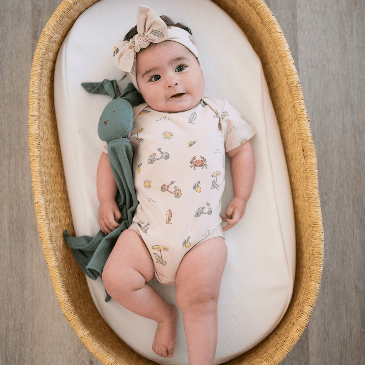 Baby girl lying in basket wearing Summer Vibes print short sleeve bodysuit with matching bow and holding a green bunny lovey
