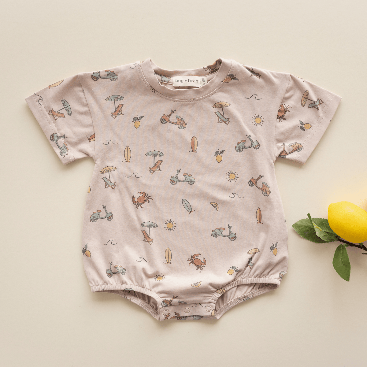 Flat lay of bug + bean Summer Vibes bubble romper with playful beach print and lemon prop on cream background