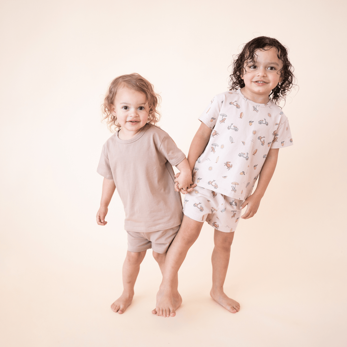 Two toddlers holding hands in matching bug + bean kids organic cotton short sets—one in Summer Vibes print and one in solid mocha, ideal for sibling style moments.
