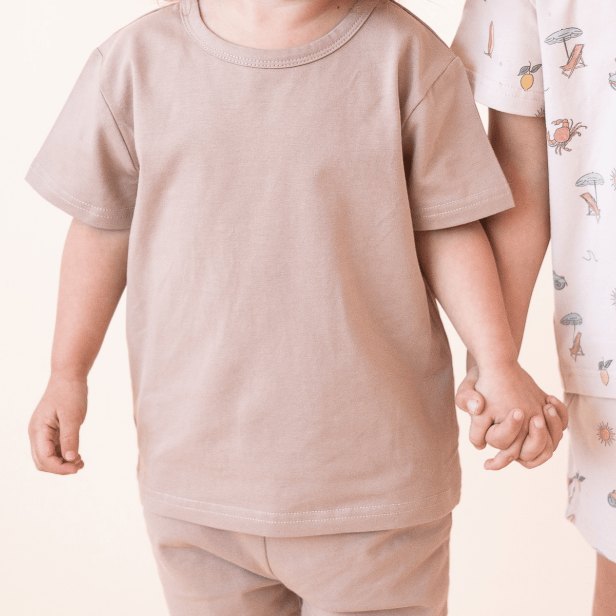 Close-up of bug + bean kids mocha organic cotton toddler short set with neutral tones, shown while two children hold hands.