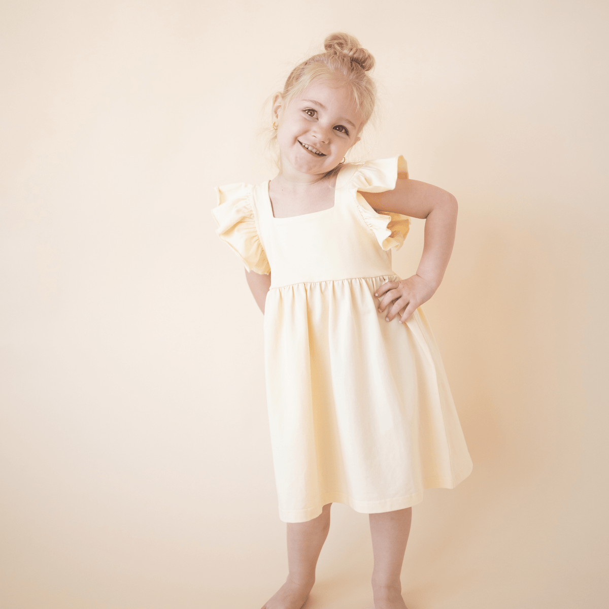 oddler girl smiling in bug + bean kids lemonade yellow organic cotton dress with ruffle sleeves, perfect for summer play or special moments.