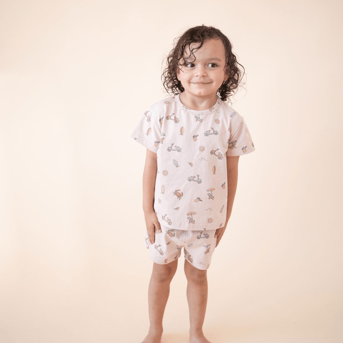 oddler boy smiling in bug + bean kids Summer Vibes organic cotton short set, featuring playful beach-themed illustrations perfect for warm-weather days.